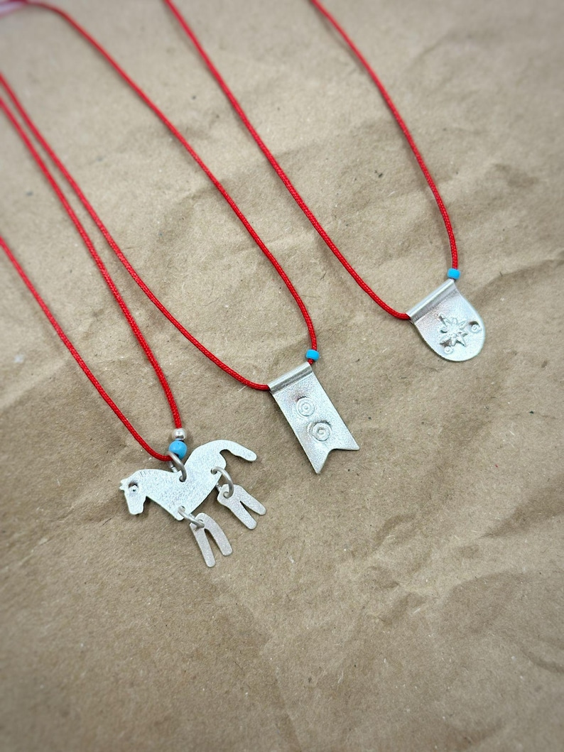 May include: Three necklaces featuring silver-toned metal pendants on thin red cords. One pendant is shaped like a horse with dangling legs, one is a rectangular flag with two circular engravings, and one is a rounded shield with a star design.