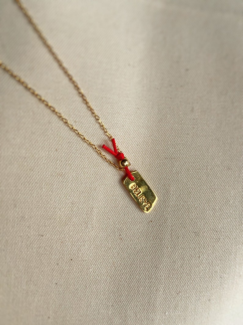 May include: A gold chain necklace with a small gold tag that says "Believe" hanging from it. The tag is attached to a red string with a gold bead.