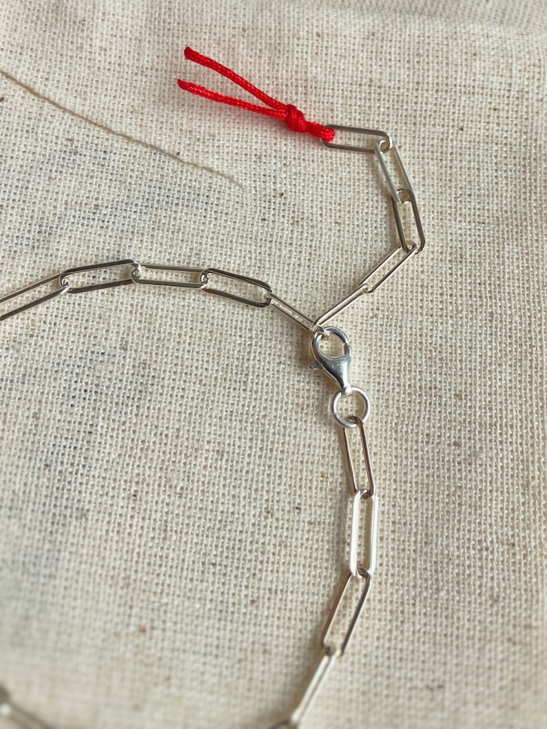 Pode incluir: Um colar de corrente de prata com fecho de lagosta. A corrente &eacute; feita de pequenos elos retangulares. Um cord&atilde;o vermelho est&aacute; preso &agrave; corrente.