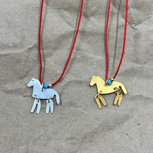 May include: Two necklaces featuring horse-shaped pendants, one in silver-tone and one in gold-tone metal. Each pendant hangs from a thin red cord accented with a small turquoise bead and a tiny metallic spacer bead near the horse figure.