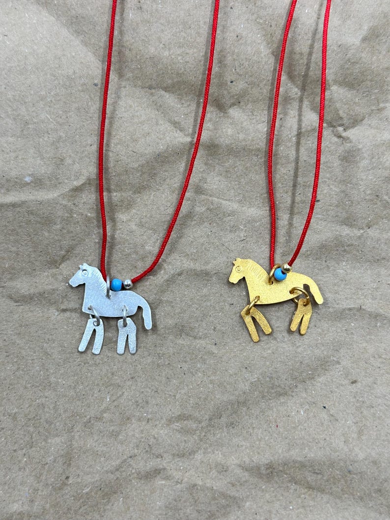 May include: Two necklaces featuring horse-shaped pendants, one in silver-tone and one in gold-tone metal. Each pendant hangs from a thin red cord accented with a small turquoise bead and a tiny metallic spacer bead near the horse figure.