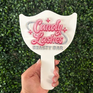 May include: A white handheld sign with a handle featuring the pink 3D text Candy Lashes surrounded by a silver ring and small star accents. Below the main text, the words BEAUTY BAR are printed in a simple, clean, silver-colored font.
