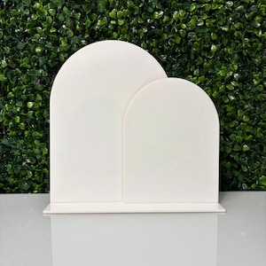 May include: Two white, arched-top display stands of different sizes, set on a white base. The stands are made of a smooth, solid material and are positioned in front of a green hedge background.