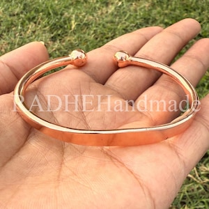 May include: A copper-colored cuff bracelet with rounded ball ends. The bracelet has a flat, polished surface and the text "RADHEHandmade" is visible on the bracelet. The bracelet is held in a hand against a background of green grass.
