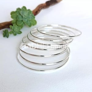 Op de afbeelding: Een stapel zilveren armbanden met een gepolijste afwerking. De armbanden liggen op een witte ondergrond, met een vetplant en een stuk drijfhout op de achtergrond. De armbanden hebben een diameter van ongeveer 6,35 cm.
