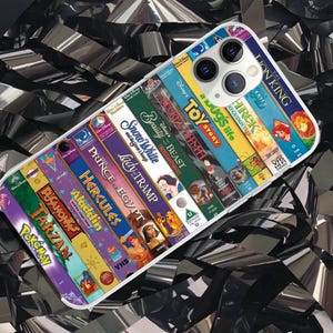 May include: A clear phone case with a design of VHS tapes. The case features titles such as "The Lion King," "Toy Story," and "Beauty and the Beast." The case has a white border and a camera cutout.