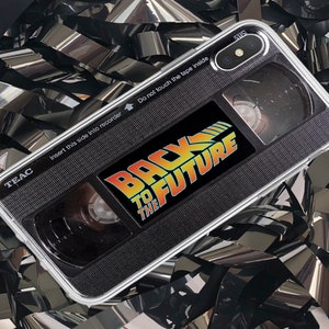 Puede incluir: Una funda transparente para teléfono con un diseño negro y plateado que parece una cinta VHS. La funda tiene las palabras "Regreso al futuro" impresas en ella.