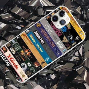 May include: A white phone case featuring a design of VHS movie covers, including "Jurassic Park," "Back to the Future," and "Star Wars." The case has a clear border and a camera cutout. The background is a shiny, black, ribbon-like material.