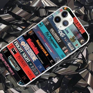 May include: A phone case displaying a collection of VHS movie covers, such as "Predator," "Halloween," and "Nightmare on Elm Street." The case has a white border and a glossy finish, with the phone's camera visible. The background is a shiny, metallic ribbon.