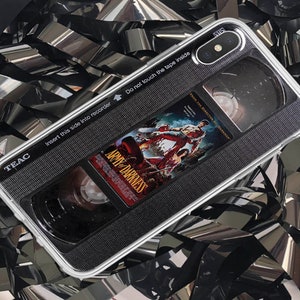 May include: A black and white phone case with a vintage VHS tape design. The case features the text "TEAC" and "Insert this side into recorder" at the top. The case also features the text "Do not touch the tape inside" and "Army of Darkness" at the bottom. The case has a clear plastic window that shows the inside of the tape.