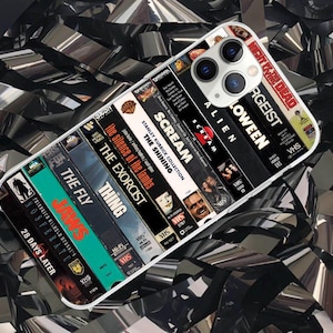 May include: A phone case featuring a design of VHS tapes. The case displays titles like "JAWS", "ALIEN", "SCREAM", and "THE SHINING". The case is white with a clear border and is set against a background of shiny, black material.
