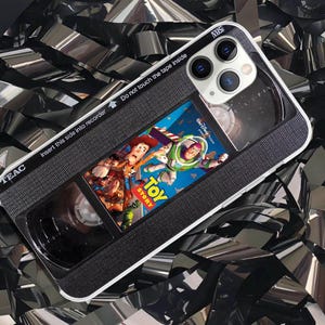 Puede incluir: Una funda de teléfono diseñada para parecerse a una cinta VHS, con la portada de la película "Toy Story". La funda es negra con ribetes blancos e incluye el texto "TEAC", "Insert this side into recorder" y "Do not touch the tape inside".