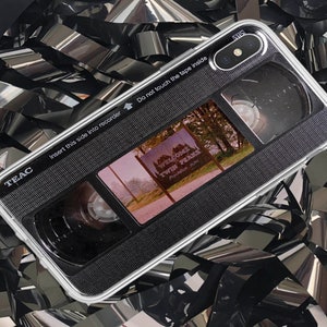 May include: A white phone case with a black cassette tape design. The case has a clear window showing a scene from the TV show "Twin Peaks" with the text "Welcome Twin Peaks" on a sign. The case also has the text "TEAC" and "Insert this side into recorder" printed on it.