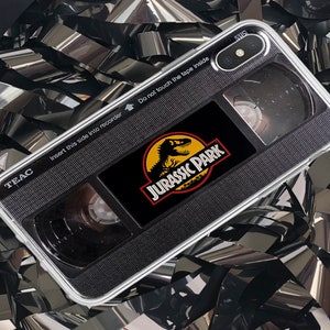 Puede incluir: Una funda para teléfono negro y plateado con un diseño de cinta VHS de Jurassic Park. La funda tiene una parte posterior transparente y una parte delantera negra con el logotipo de Jurassic Park y una silueta de dinosaurio. El texto "TEAC", "Inserte este lado en la grabadora" y "No toque la cinta en el interior" está impreso en la funda.