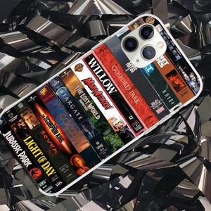 Op de afbeelding: Een smartphonehoesje met een collage van diverse vintage VHS-filmruggen, waaronder titels zoals Jurassic Park, Light of Day, The Scorpion King, Stargate, The Lost World, Mars Attacks, Willow, Gosford Park, The Abyss en E.T.