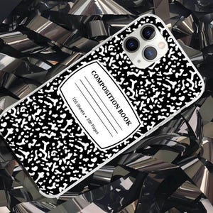 May include: A smartphone case featuring a classic black and white speckled composition notebook pattern. The design includes a central white label with the text COMPOSITION BOOK, 100 Sheets, and 200 Pages printed in black, with a camera cutout.