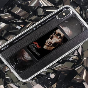 May include: A clear phone case with a black and white VHS tape design. The tape has the text "Evil Dead" and a skull graphic. The case has the text "TEAC" and "Insert this side into recorder. Do not touch the tape inside."