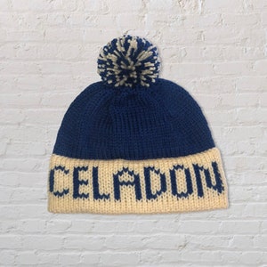 Puede incluir: Un gorro azul de punto con un pompón blanco y la palabra "CELADON" tejida en azul en el puño.