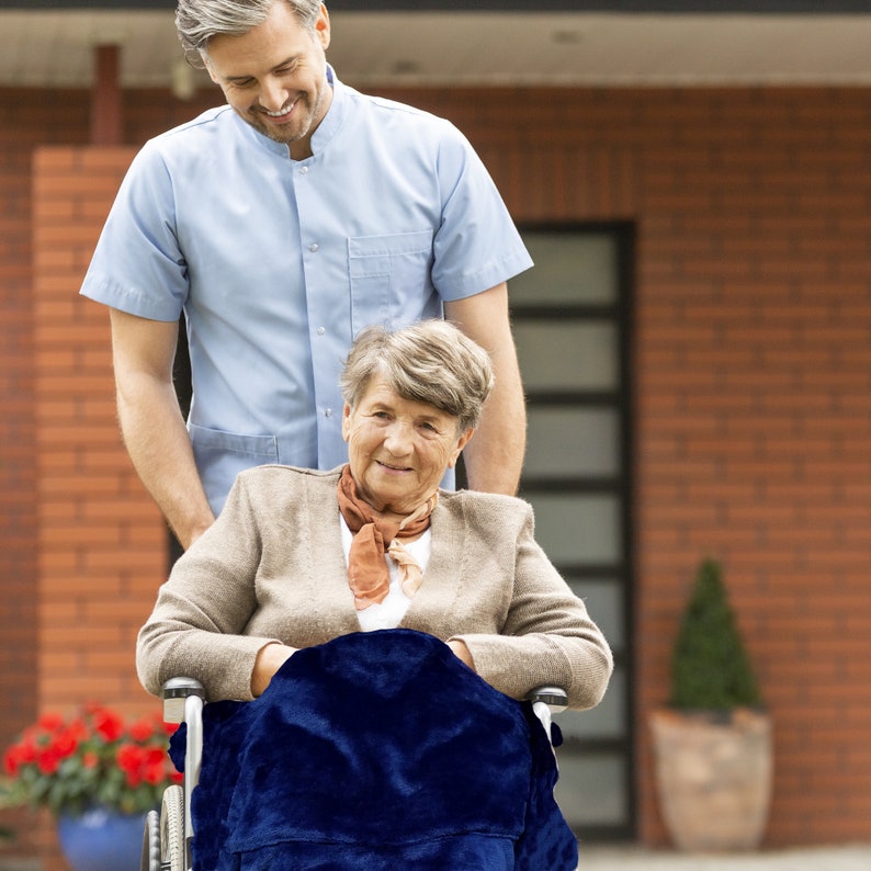 Puede incluir: Un hombre con una camisa azul claro ayuda a una mujer mayor en una silla de ruedas. La mujer lleva una chaqueta marr&oacute;n y una manta azul. Ella sonr&iacute;e y mira a la c&aacute;mara.