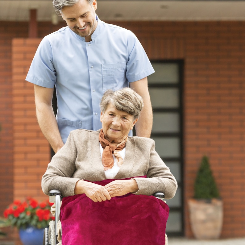 May include: A woman in a wheelchair smiles at a man in a light blue shirt. The woman is wearing a brown jacket and a red scarf. The wheelchair has a red blanket on the seat.