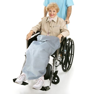 May include: A senior woman in a wheelchair is being pushed by a person wearing a light blue shirt. The woman is wearing a beige jacket and a blue blanket. The wheelchair has black wheels and a black seat.
