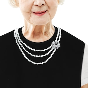 May include: A woman wearing a black dress with a three-strand pearl necklace and a silver rose brooch.