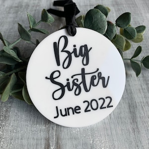 May include: A white circular ornament with the words "Big Sister June 2022" in black script. The ornament has a black ribbon for hanging and is surrounded by green foliage.
