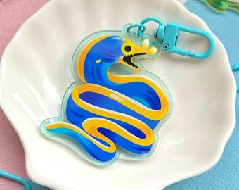 May include: A collection of colorful acrylic keychains featuring marine life. The central keychain is a blue and yellow moray eel. Other keychains include fish and a stingray, each with a metal clasp. The keychains are displayed on a white seashell-shaped dish.