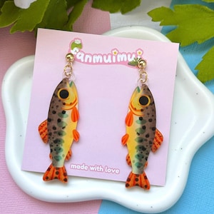 May include: A pair of dangle earrings featuring a fish design. The earrings have a yellow body with brown spots, orange fins, and a red tail. The earrings are hanging from gold-colored hooks. The earrings are displayed on a pink card that says "fanmuimu" and "made with love".