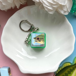以下が含まれることがあります： 魚のデザインのキーホルダー。1つのキーホルダーは、魚のイラストが入った緑色の立方体で、シルバーのキーリングが付いています。もう1つのキーホルダーは、魚の形をしたチャームが付いています。キーホルダーは、白い貝殻型の皿の上にあり、背景には白い花があります。