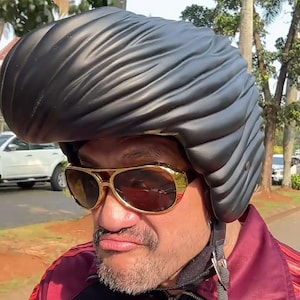 Può includere: Un casco da moto nero opaco caratterizzato da un design scolpito a forma di voluminosa acconciatura pompadour. Il casco è abbinato a occhiali da sole con montatura dorata e lenti scure, indossati sopra una giacca bordeaux e nera con zip e strisce rosse.