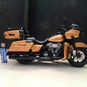 Może przedstawiać: Model motocykla w kolorze metalicznej miedzi i czerni, wyposażony w owiewkę, kufry boczne, kufer centralny z oparciem oraz czarny blok silnika. Obok motocykla umieszczono niebieską plastikową zapalniczkę dla porównania skali.