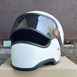 Può includere: Un casco da moto integrale bianco lucido con visiera scura oscurata, bordo in gomma nera lungo la base e l'apertura oculare, e inserti laterali in pelle marrone fissati con rivetti metallici color oro su entrambi i lati della calotta.