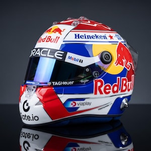 Può includere: Un casco da corsa con una combinazione di colori bianco, blu e rosso e il marchio Red Bull. La visiera mostra i loghi Oracle, Mobil 1 e TAG Heuer. Sono presenti anche i marchi Heineken, viaplay, Gate.io e il testo Verstappen.com.