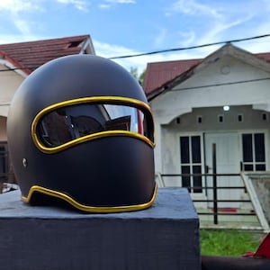 May include: A matte black motorcycle helmet featuring a horizontal visor opening outlined with a bright gold trim. The helmet has a smooth, rounded shell design with a gold-trimmed lower edge, providing a sleek and uniform appearance throughout.