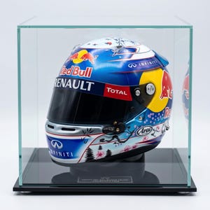 Può includere: Un casco Red Bull Racing racchiuso in una teca di vetro trasparente. Il casco presenta una base blu e bianca con loghi Red Bull, grafiche di fiori di ciliegio e marchi Infiniti, Renault e Total. Una visiera nera copre la parte anteriore.