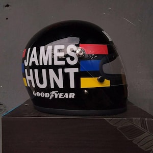 Può includere: Un casco da corsa nero con il nome JAMES HUNT in grandi lettere maiuscole bianche sul lato. Strisce orizzontali rosse, blu e gialle corrono lungo il casco e il logo Goodyear è stampato in bianco vicino al bordo inferiore.
