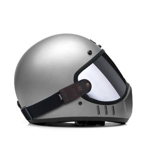 May include: A matte silver full-face helmet featuring a dark tinted visor, a black elastic strap with a brown leather-like side accent, and three vertical ventilation slits on the chin guard. The word TROOPER is printed on the black strap.