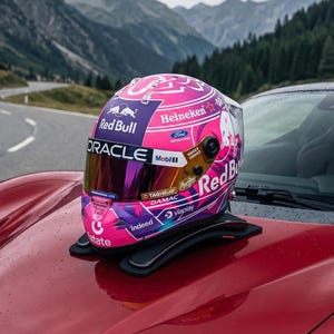 Puede incluir: Un casco de carreras rosa y morado con logotipos de Red Bull, Oracle, Heineken, Ford, Mobil 1, Tag Heuer, Damac, Indeed y Viaplay. El casco tiene una visera reflectante y está colocado sobre un soporte negro en el capó de un vehículo rojo.