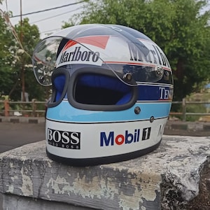 Può includere: Un casco integrale bianco, blu e rosso con visiera trasparente e loghi ben visibili come Marlboro, BOSS e Mobil 1. Il casco presenta una calotta arrotondata con bordo nero attorno all'apertura e un'imbottitura interna di colore blu.