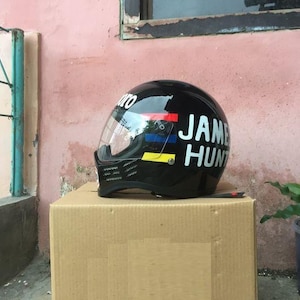 Può includere: Un casco integrale da moto nero dotato di visiera trasparente e mentoniera con feritoie di ventilazione. Il lato del casco riporta la scritta "JAMES HUNT" in lettere maiuscole bianche, accompagnata da strisce orizzontali rosse, blu e gialle.