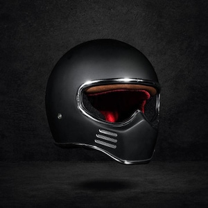 Può includere: Un casco da moto integrale nero opaco caratterizzato da un'apertura della visiera con bordo cromato, una fodera interna in similpelle marrone e tre feritoie di ventilazione cromate orizzontali sulla mentoniera del casco.