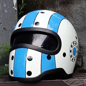 May include: A white motorcycle helmet featuring vertical light blue stripes and black circular polka dots. The helmet includes a wide, horizontal black visor opening and a circular graphic on the side resembling a vintage rotary telephone dial.