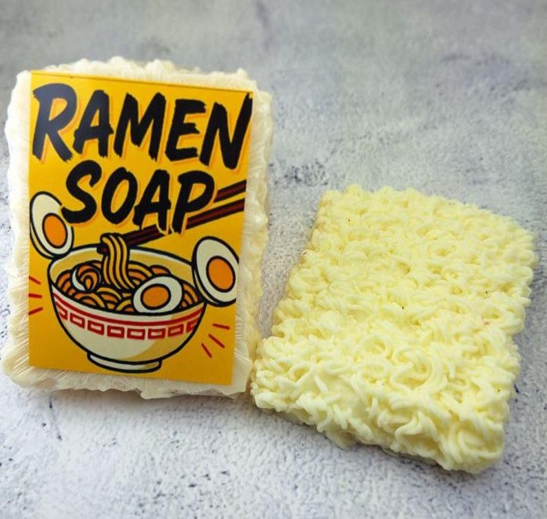 May include: Two bars of soap, one with a yellow label that reads "RAMEN SOAP" with a graphic of ramen noodles and eggs. The other bar is shaped like ramen noodles and is a light yellow color.