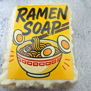 May include: A bar of soap with a yellow label featuring the words "RAMEN SOAP" in black. The label has a cartoon illustration of a ramen bowl with noodles, eggs, and chopsticks. The soap itself is a light beige color.