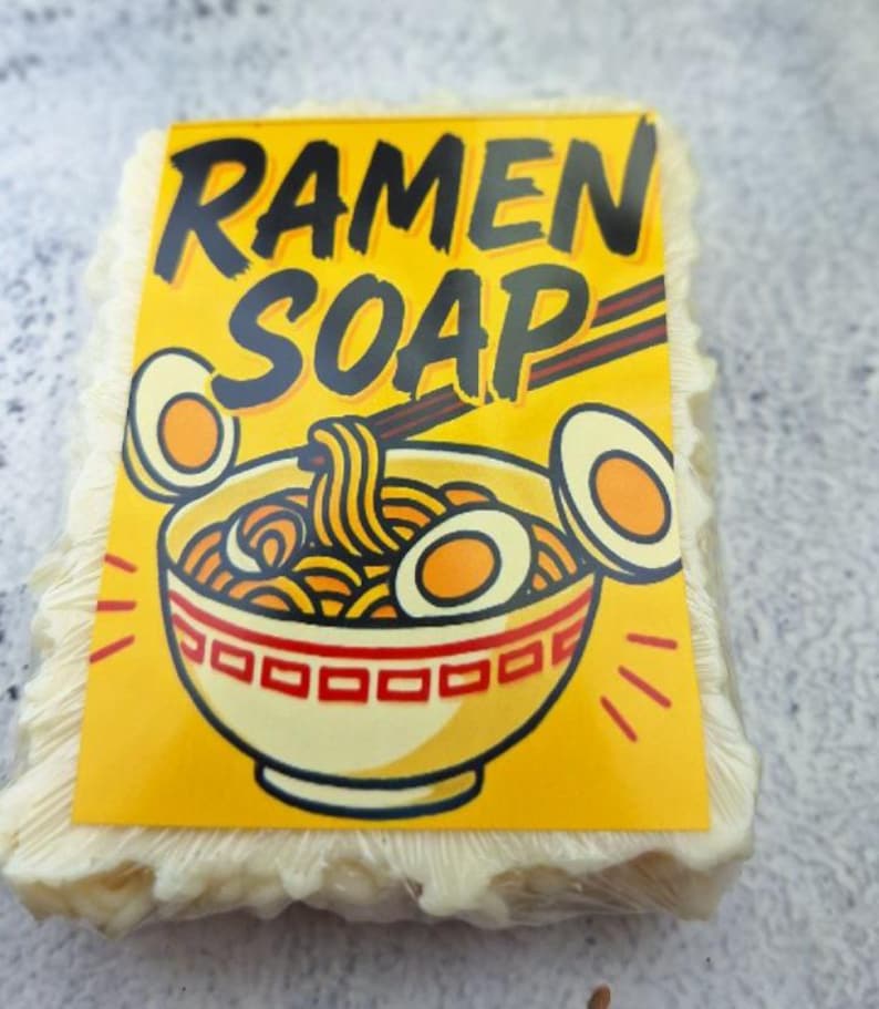 May include: A bar of soap with a yellow label featuring the words "RAMEN SOAP" in black. The label has a cartoon illustration of a ramen bowl with noodles, eggs, and chopsticks. The soap itself is a light beige color.