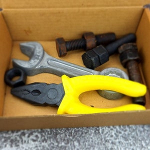 May include: A collection of tools, including a wrench, pliers with yellow handles, nuts, and bolts, are arranged inside a cardboard box. The tools are in various shades of gray, black, and brown.