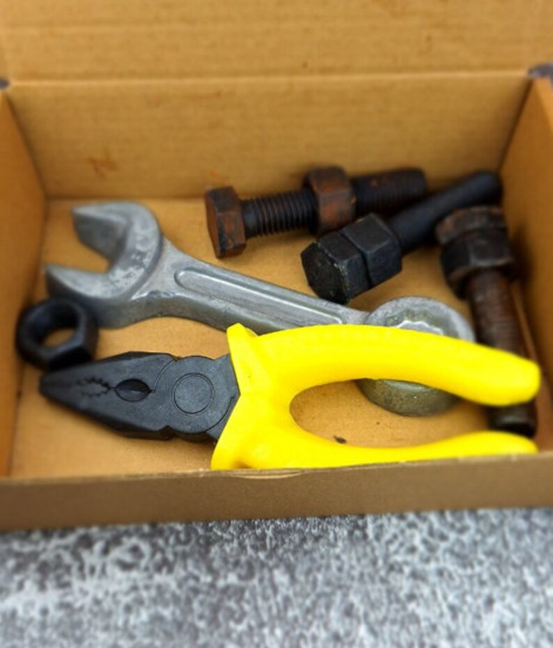 May include: A collection of tools, including a wrench, pliers with yellow handles, nuts, and bolts, are arranged inside a cardboard box. The tools are in various shades of gray, black, and brown.