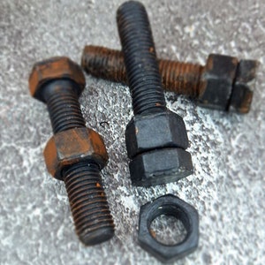 May include: Close-up of several black bolts and nuts with visible rust. The bolts have hexagonal heads and threaded shafts. One nut is separate from the bolts. The objects are on a textured gray surface.