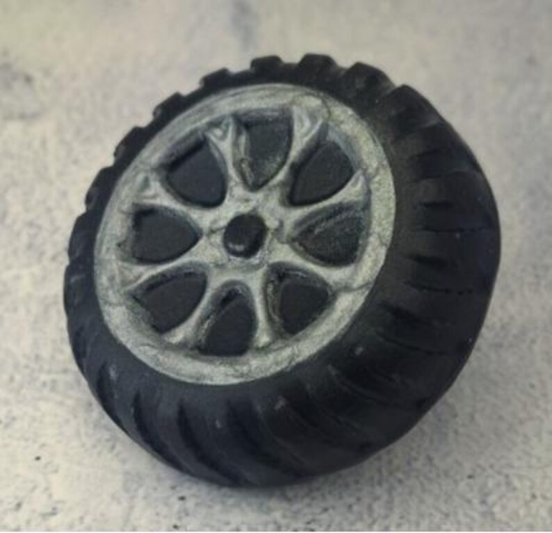 May include: A miniature black tire with a silver rim. The tire has a detailed tread pattern and a multi-spoke wheel design. The wheel is a detailed replica of a car or truck tire.